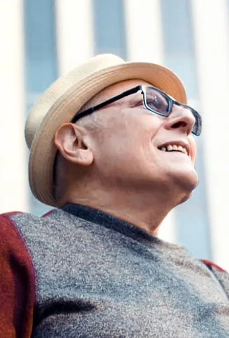 Michael Chanak Jr., smiling and looking up, in a straw hat, glasses and a gray sweater with red sleeves, photographed from below against a tall building.